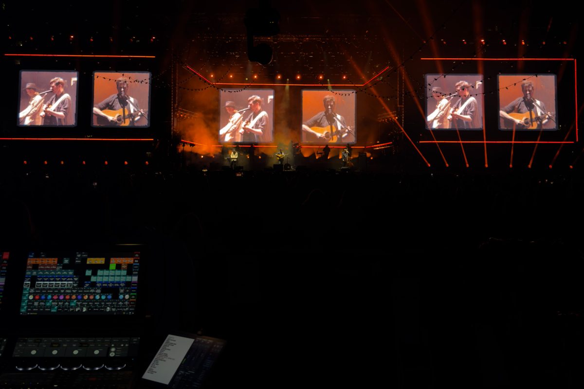 Front-of-house view of a Mumford and Sons show, the lighting console in the foreground and the band on the stage LED screens